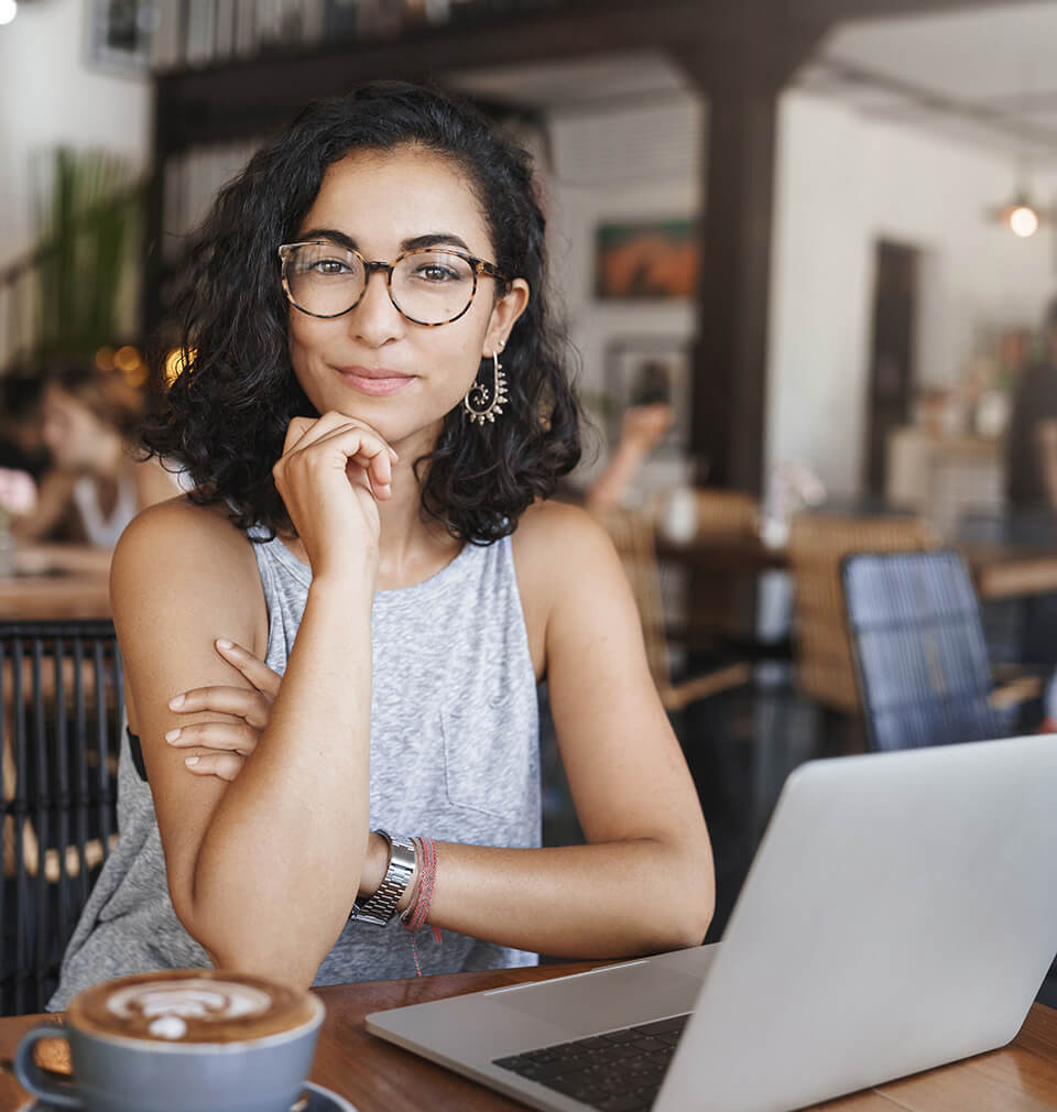 a woman looks directly at the camera with coffee and laptop in front of her