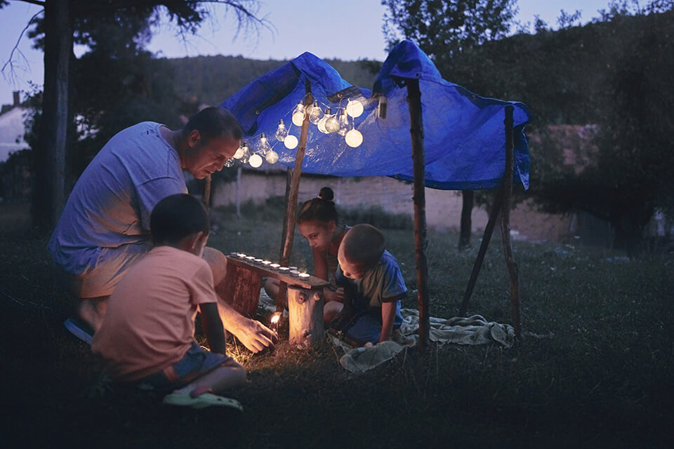 a father lights a candle with his kids under a blue tarp